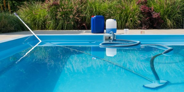 The image showcases a clear blue swimming pool, with a cleaning hose submerged in the water. Chemical containers and a water testing kit are positioned on the poolside, highlighting the focus on maintaining optimal water quality. The surrounding lush greenery adds to the serene and well-kept atmosphere, emphasizing the importance of regular pool care and maintenance.